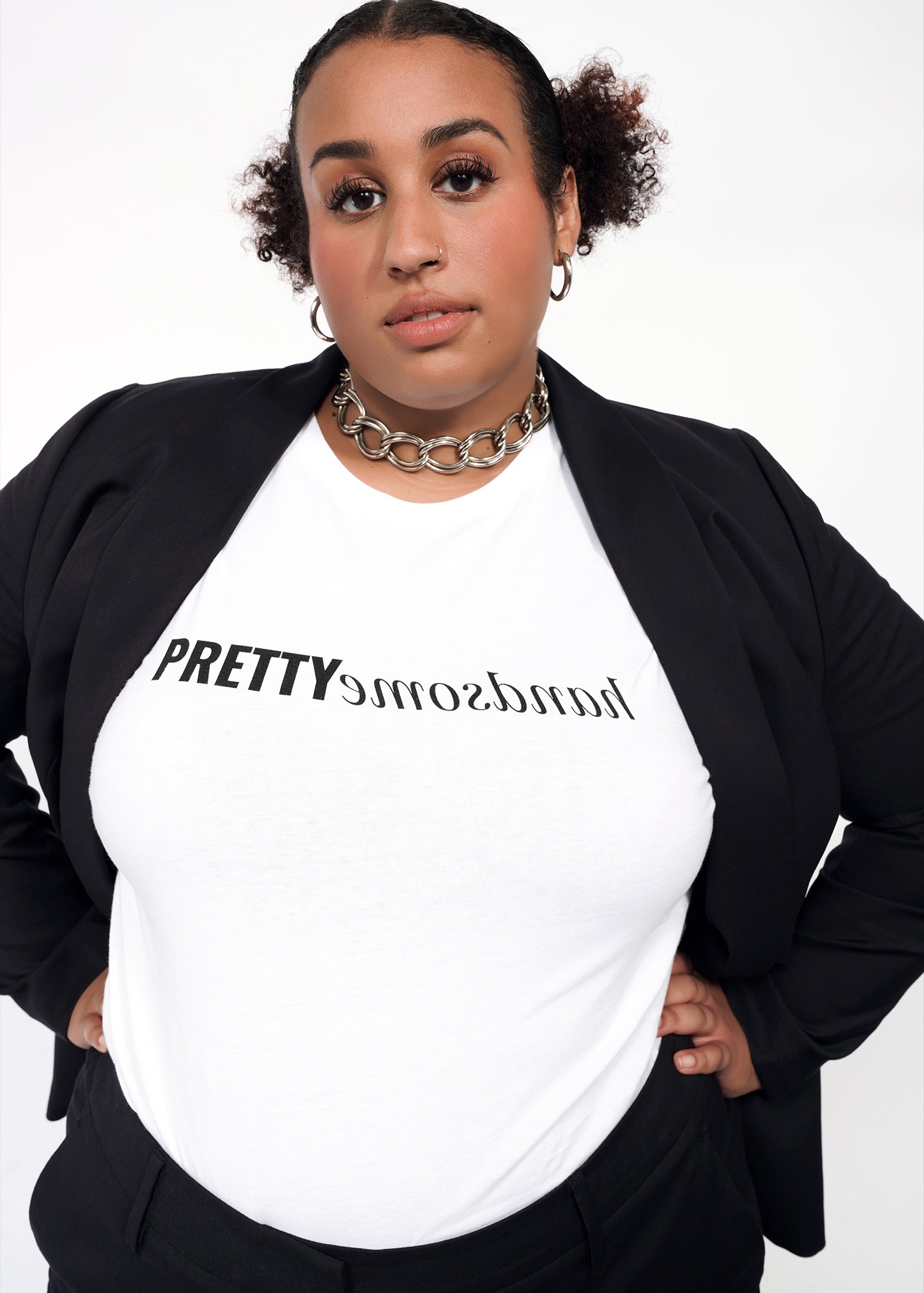 1X=A person wearing a white crew neck t-shirt with the text 'PRETTY handsomE' on the center chest area, paired with a black blazer and trousers, accessorized with a necklace.