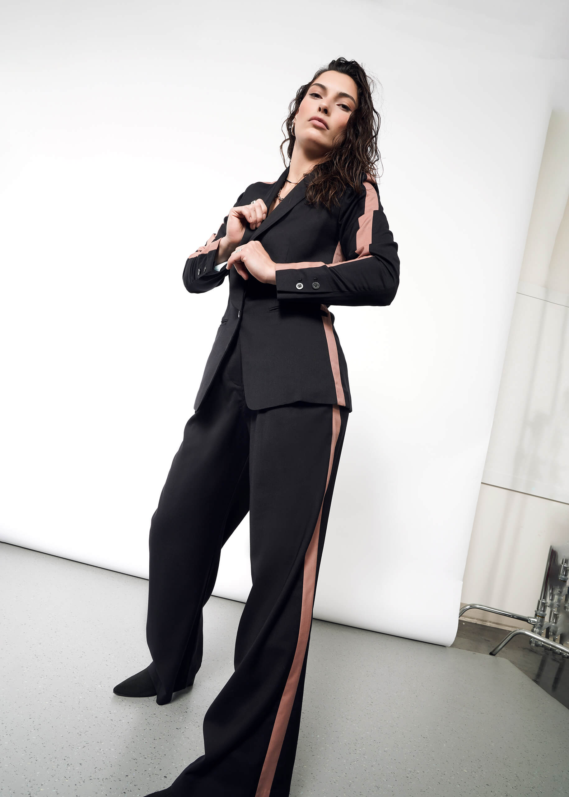 S=A woman confidently poses, adjusting the buttons on her Empower Colorblock Tux Blazer with pink tuxedo stripes. The simple background highlights the blazers tailored fit.