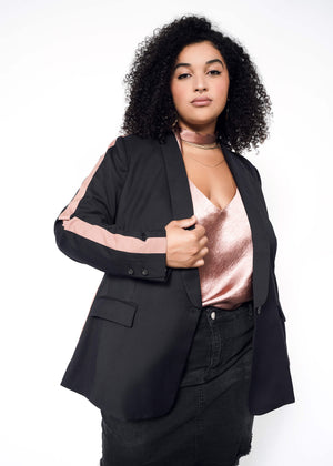1X=A woman with curly hair stands confidently against a white background, wearing The Empower Colorblock Tux Blazer—tailored with pink stripes—over a shiny pink top and black skirt.