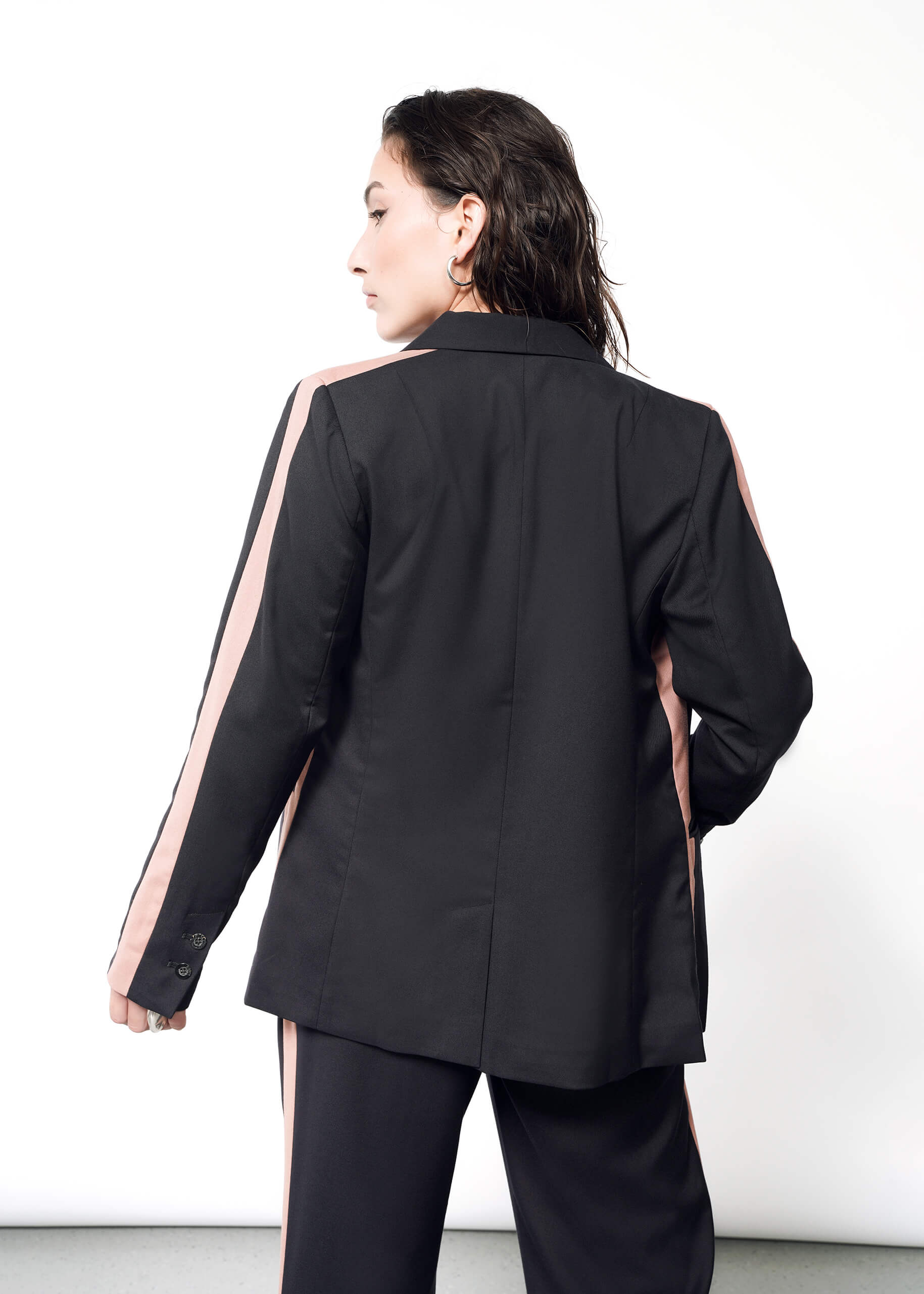 S=A person with dark hair stands against a white backdrop, wearing a stylish black suit featuring pink tuxedo stripes on the sleeves and pants. With a tailored fit, the ensemble includes The Empower Colorblock Tux Blazer, exuding sophistication and modern elegance.