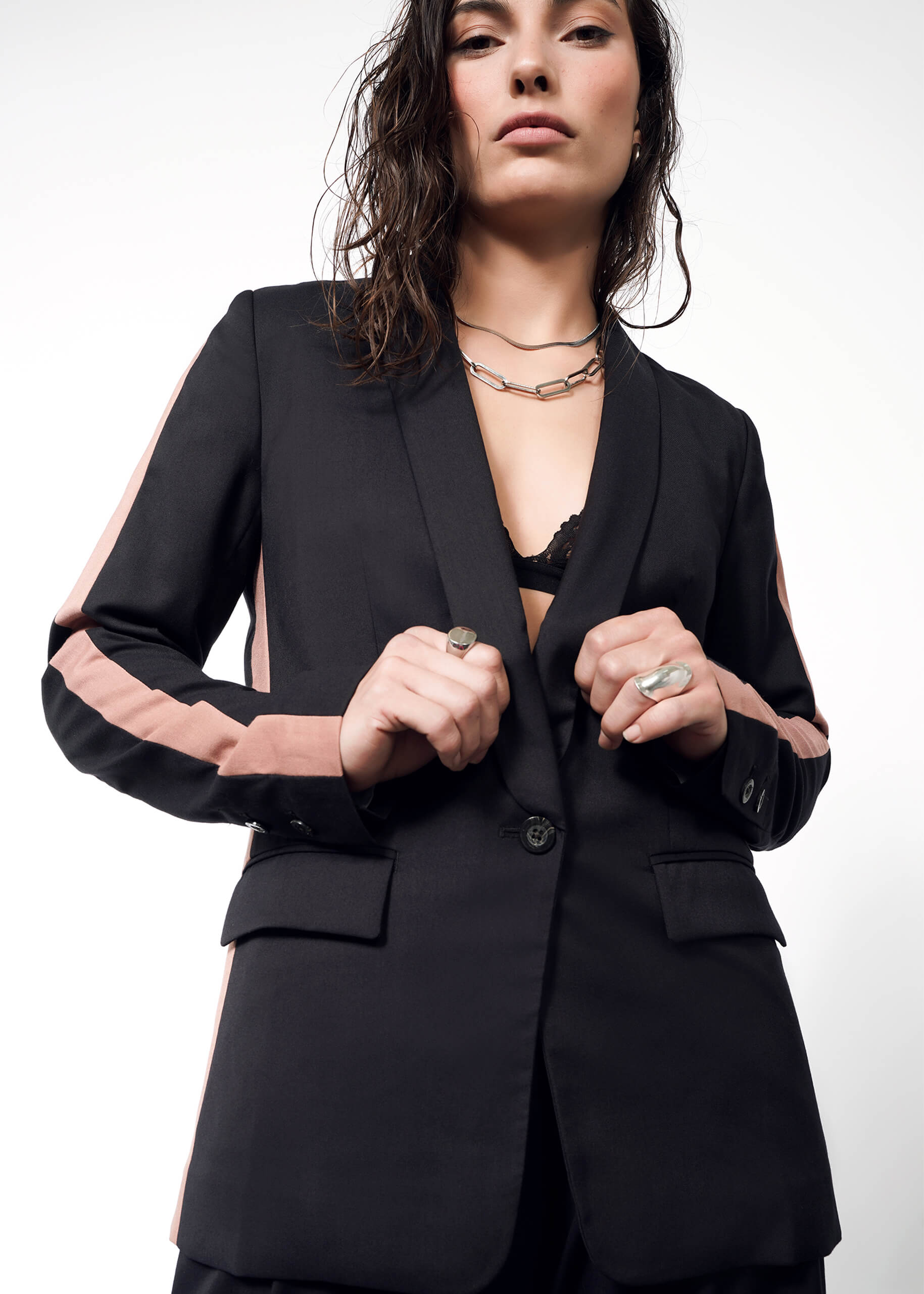 S=A woman poses confidently in The Empower Colorblock Tux Blazer, featuring a black and pink design with a deep neckline and tuxedo stripe. She wears a necklace and rings, hair loose, while gazing down at the camera against a plain white background.