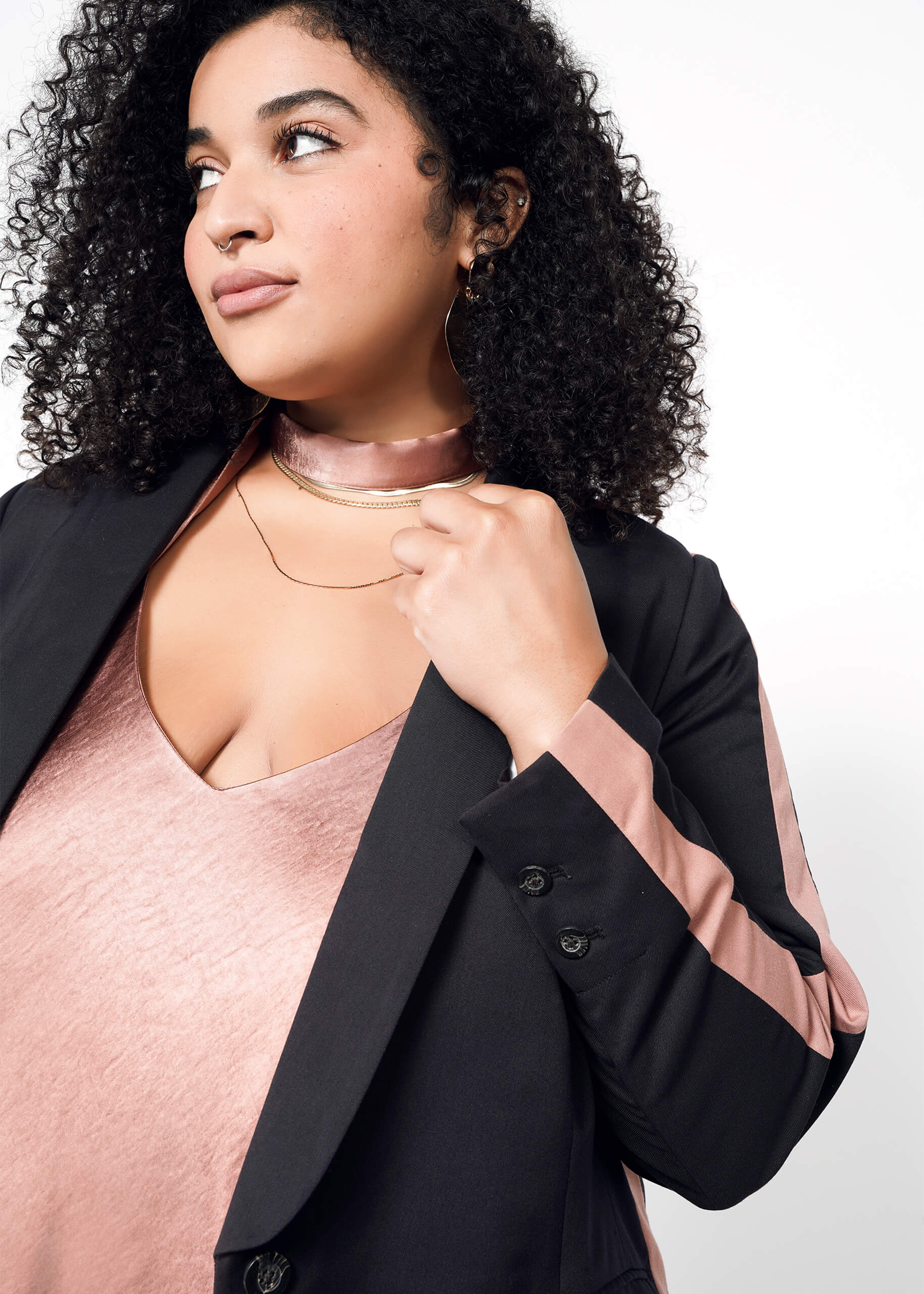 1X=A woman with curly hair looks to the side, wearing The Empower Colorblock Tux Blazer in a tailored fit with pink striped sleeves, paired with a pink satin top, choker, and layered gold necklaces against a plain white background.