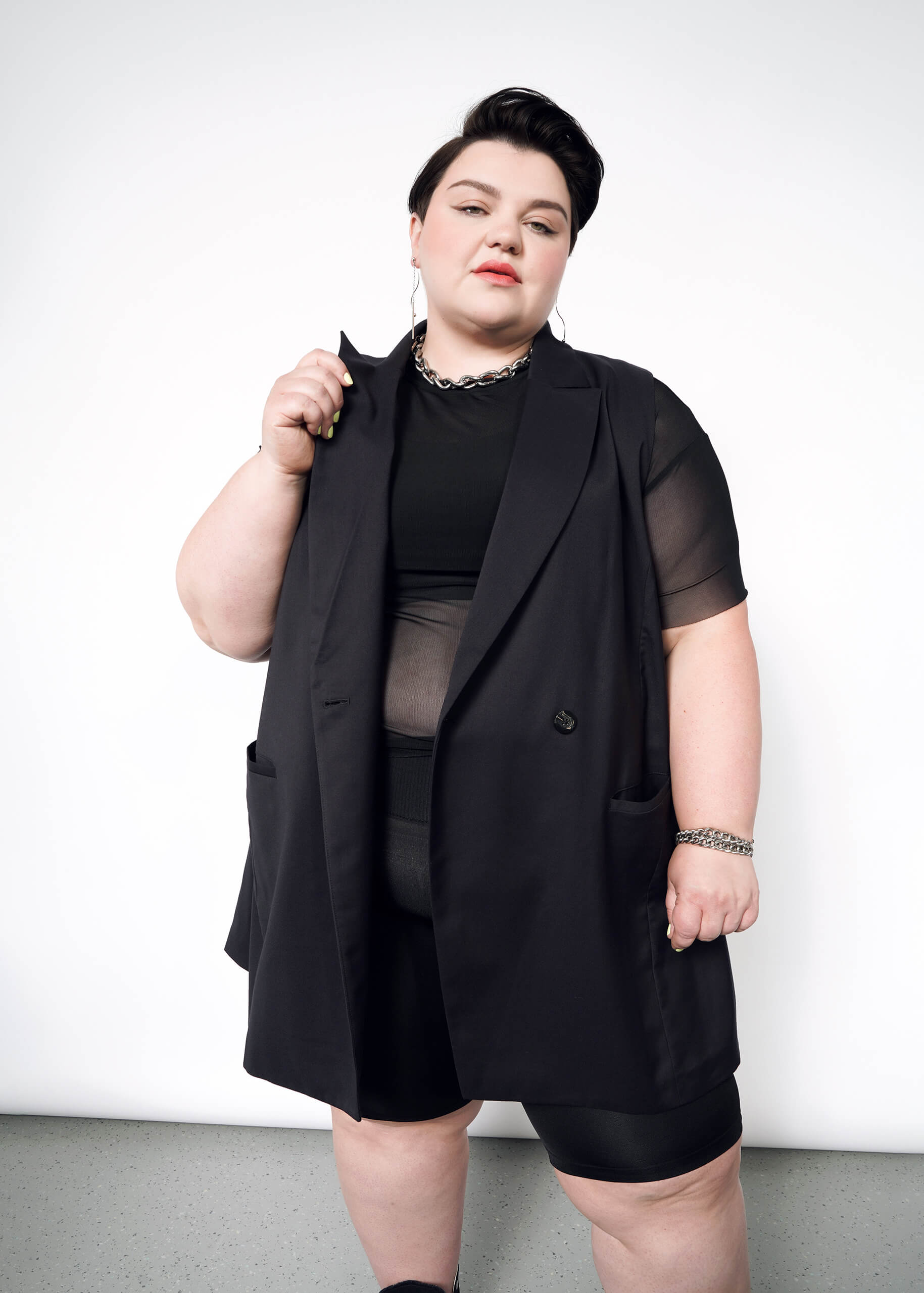1X=A person with short dark hair stands confidently against a white background, wearing The Empower Double Breasted Vest in a stylish black outfit that features a sleeveless blazer and sheer sleeve top. They hold the lapel of the oversized blazer and accessorize with silver jewelry.