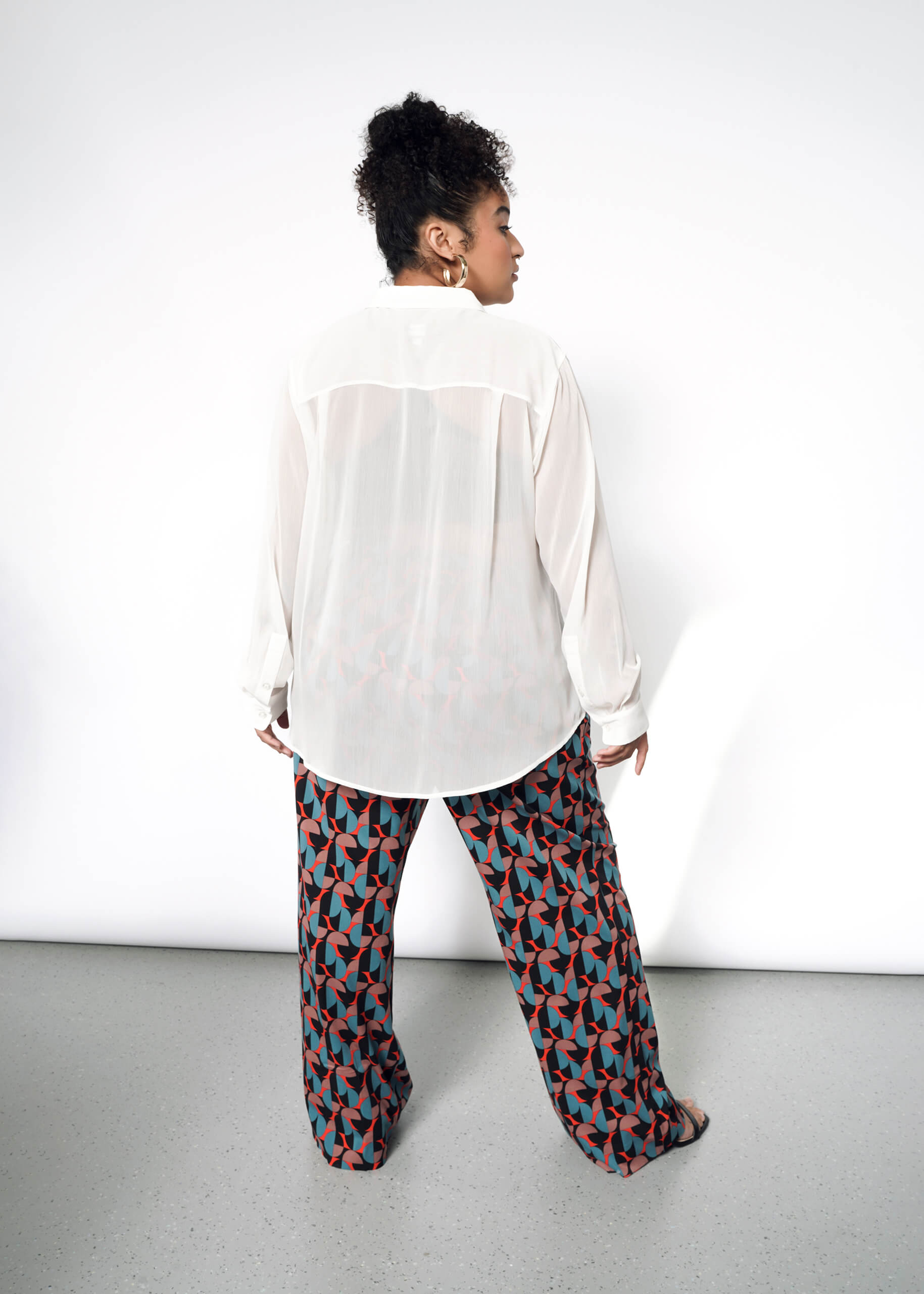 1X= A person faces away from the camera, wearing The Empower Sheer Long Sleeve Button Up and loose, wide-leg pants with a red, teal, and black geometric pattern. The background is plain white.