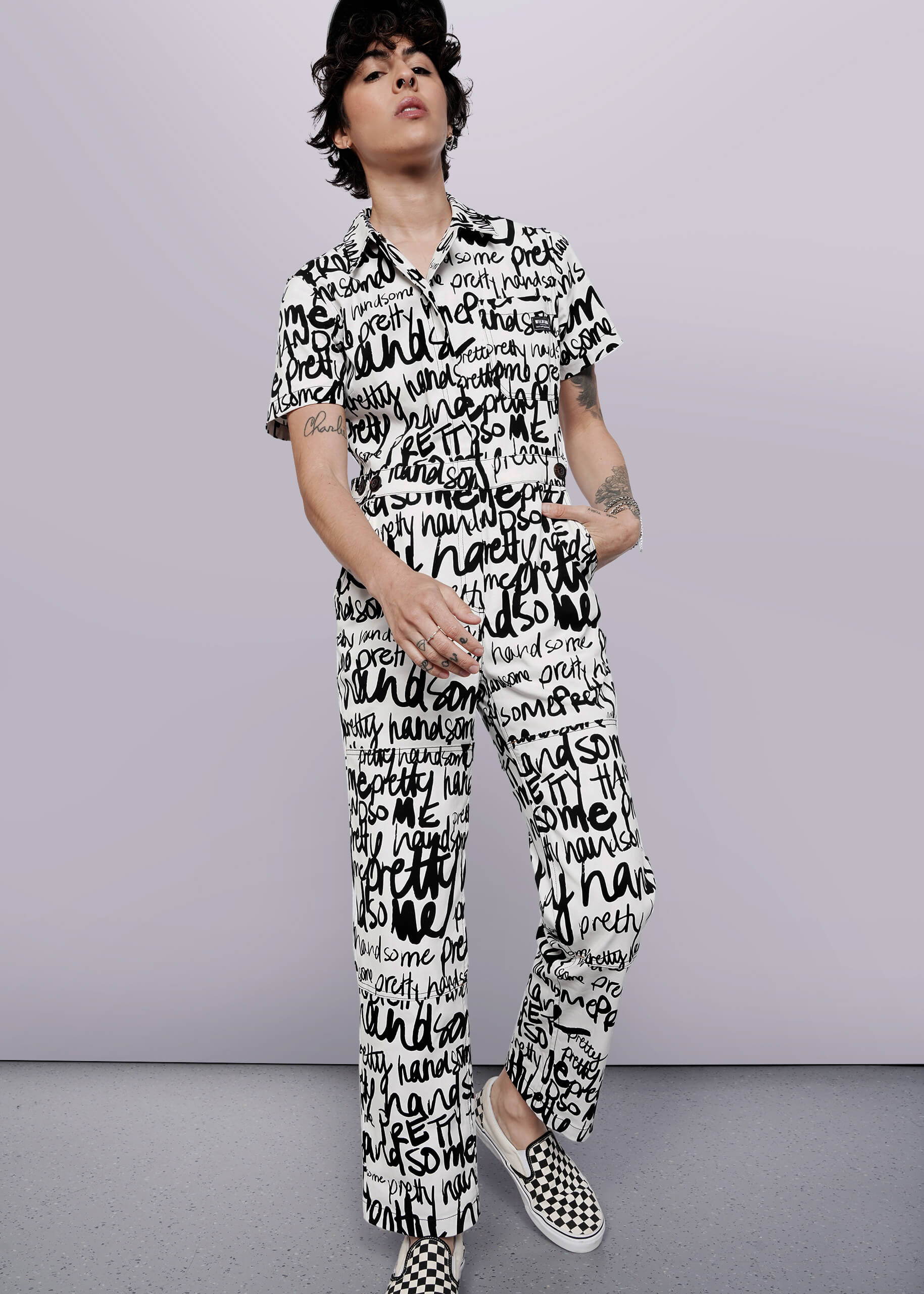 S=A person with short dark hair and arm tattoos wears The Essential High Waisted Coverall—a white jumpsuit with black handwritten words—paired with black-and-white checkered slip-on shoes, standing against a plain white background.