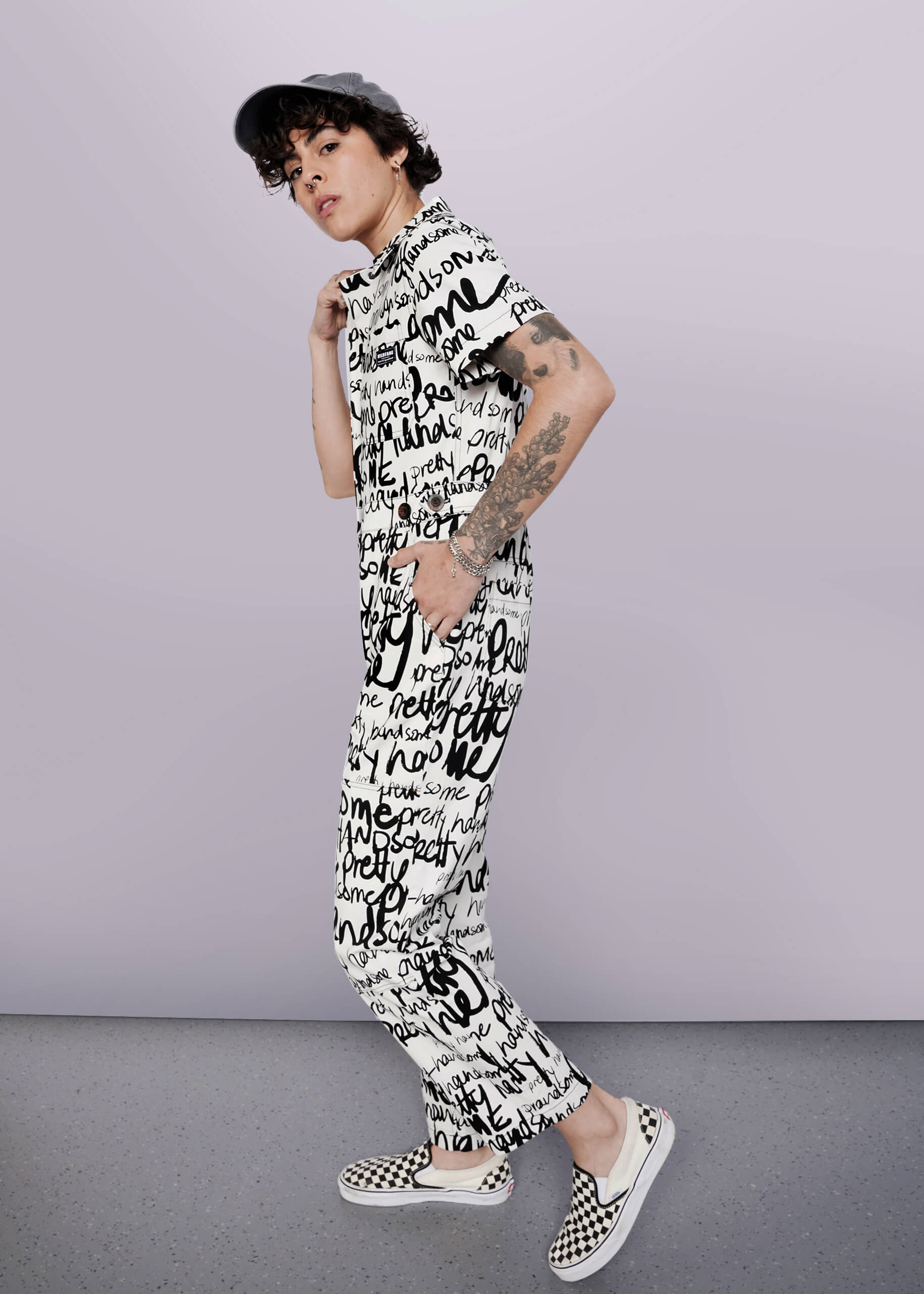 S=A person with curly short hair and visible arm tattoos poses against a white background, wearing The Essential High Waisted Coverall featuring a black and white handwritten text print, paired with a cap and checkered slip-on shoes.