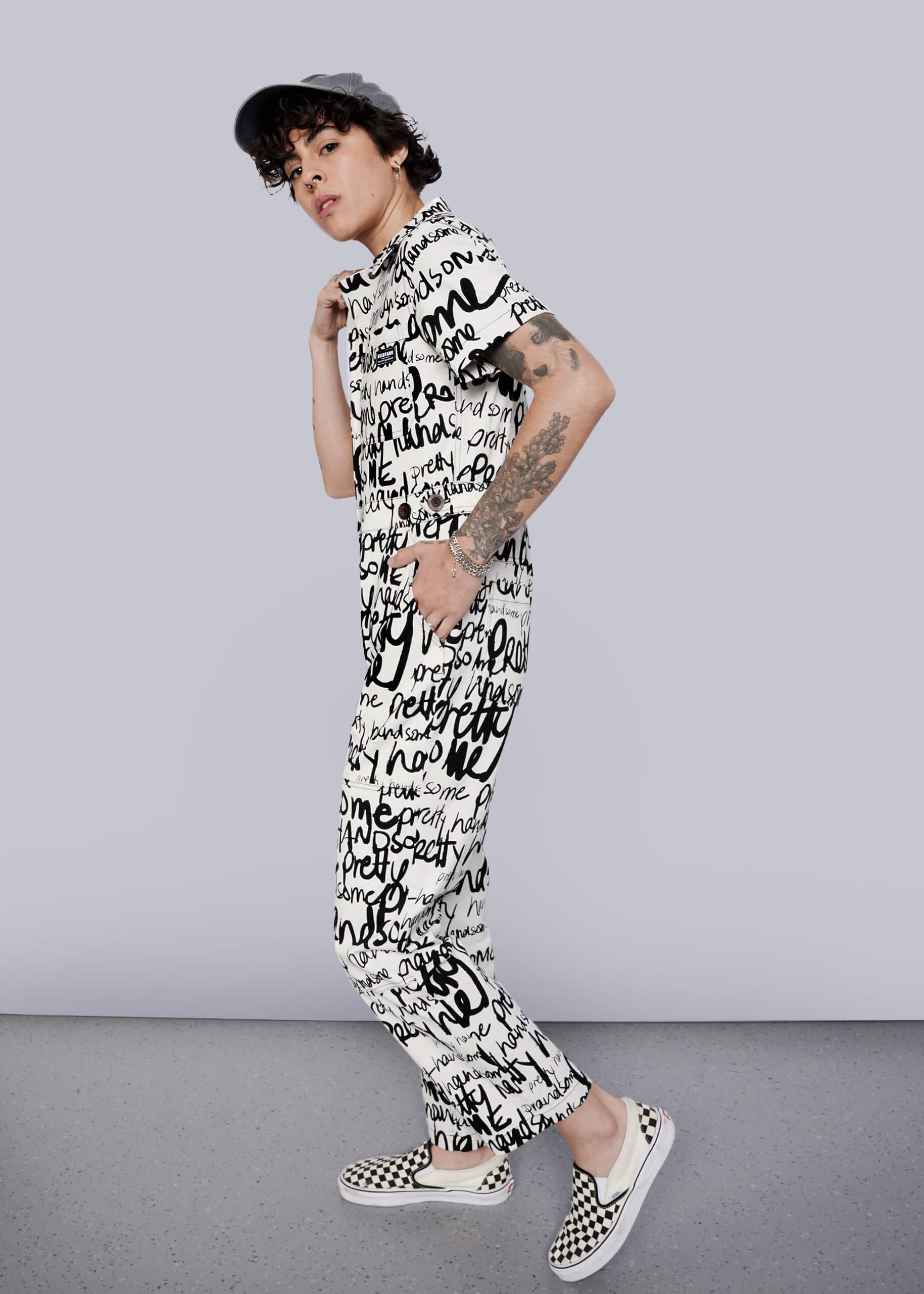 S=A person with curly short hair and visible arm tattoos poses against a white background, wearing The Essential High Waisted Coverall featuring a black and white handwritten text print, paired with a cap and checkered slip-on shoes.