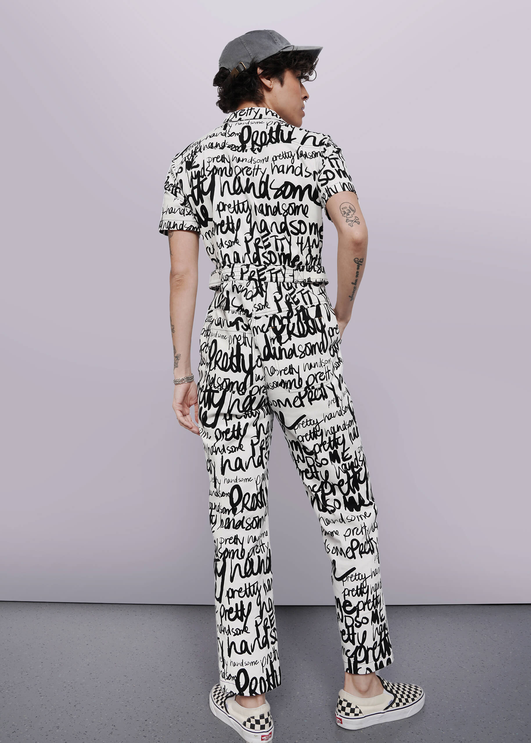 S=A person stands facing away, wearing The Essential High Waisted Coverall—a black-and-white jumpsuit with handwritten words—plus black-and-white checkerboard slip-on shoes and a gray cap. The background is plain and light-colored.