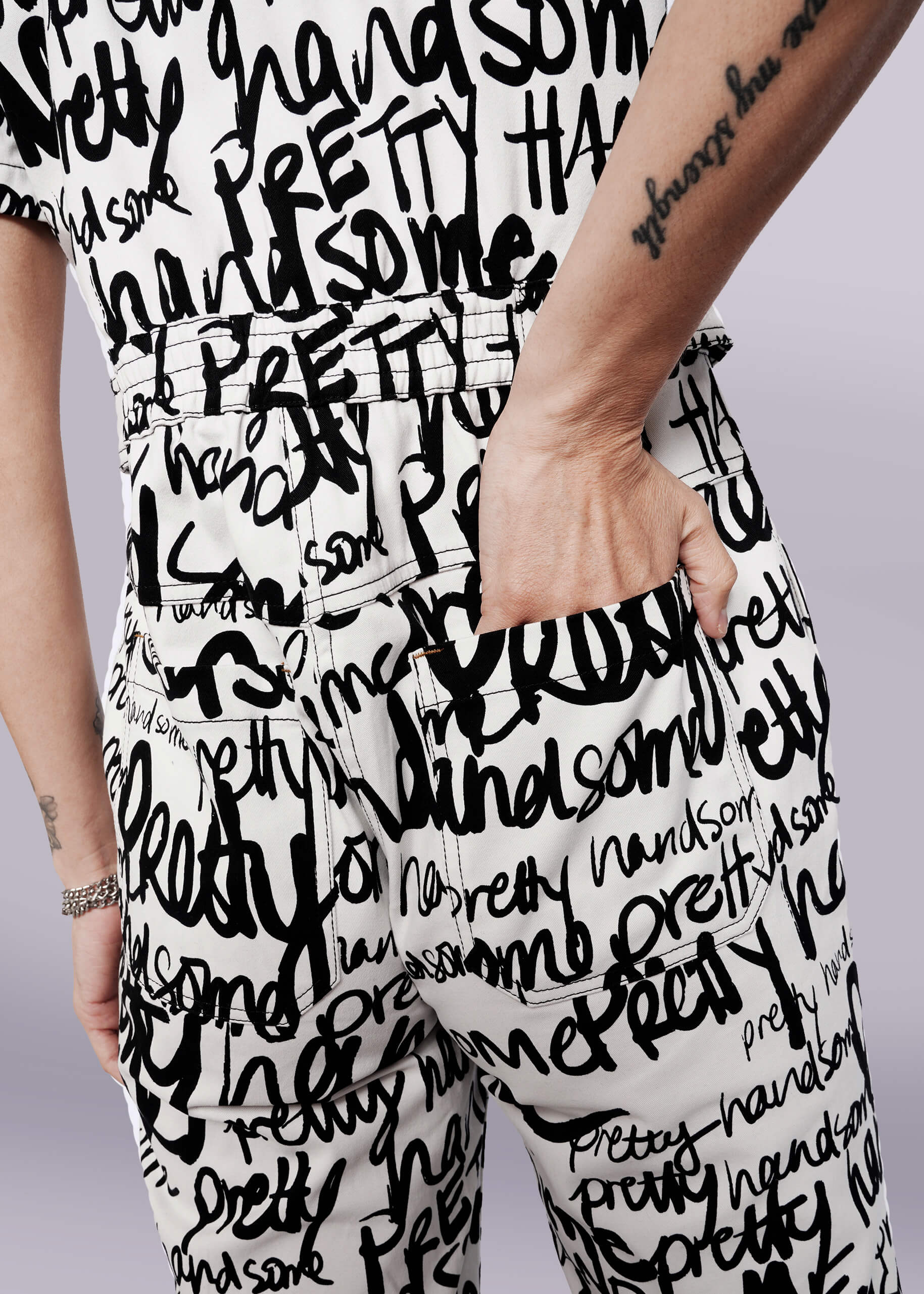 S=Wearing The Essential High Waisted Coverall, a person stands with one hand in a deep pocket; the white coverall features bold black handwritten words like pretty and handsome, and a forearm tattoo is visible.