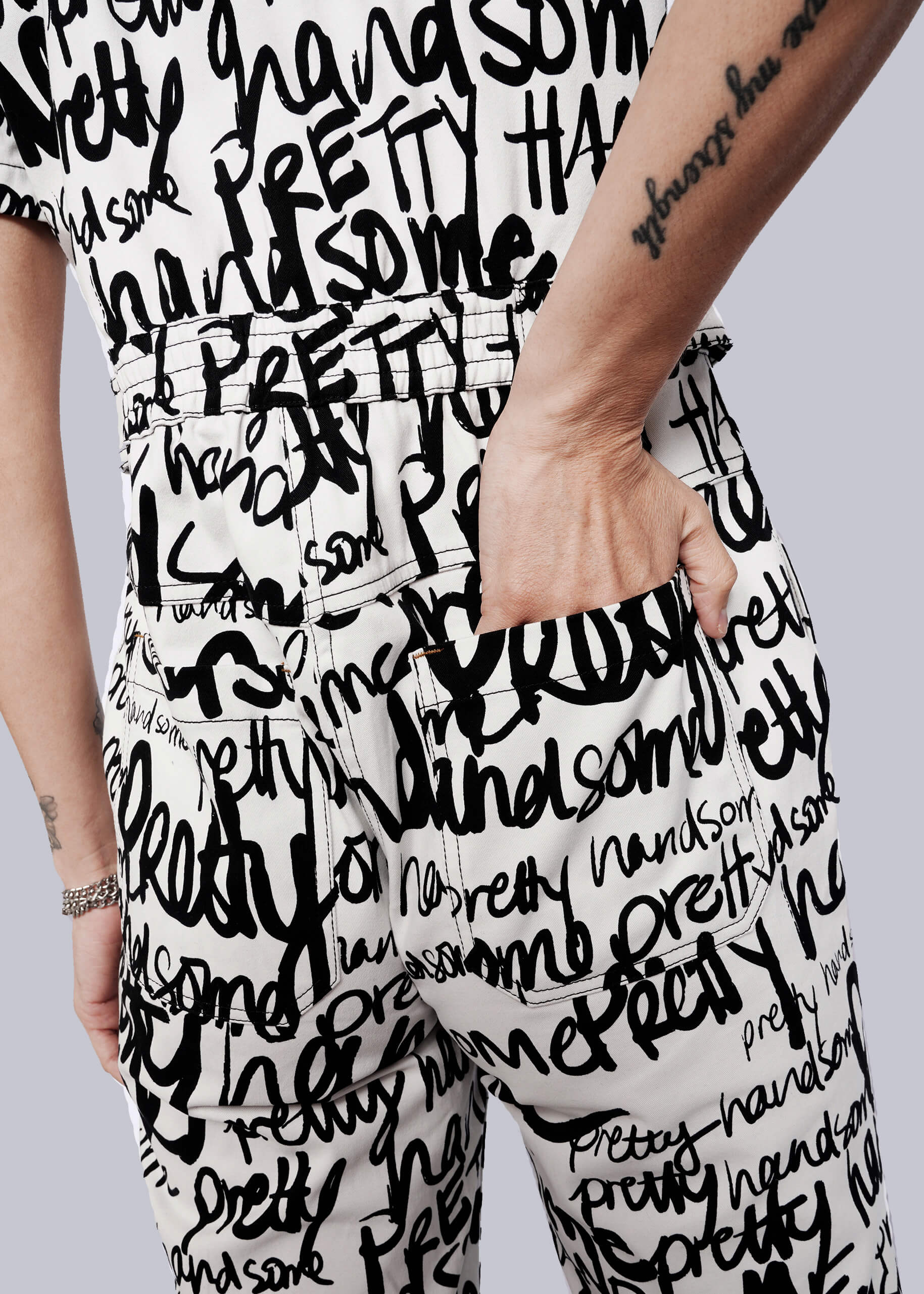 S=Wearing The Essential High Waisted Coverall, a person stands with one hand in a deep pocket; the white coverall features bold black handwritten words like pretty and handsome, and a forearm tattoo is visible.