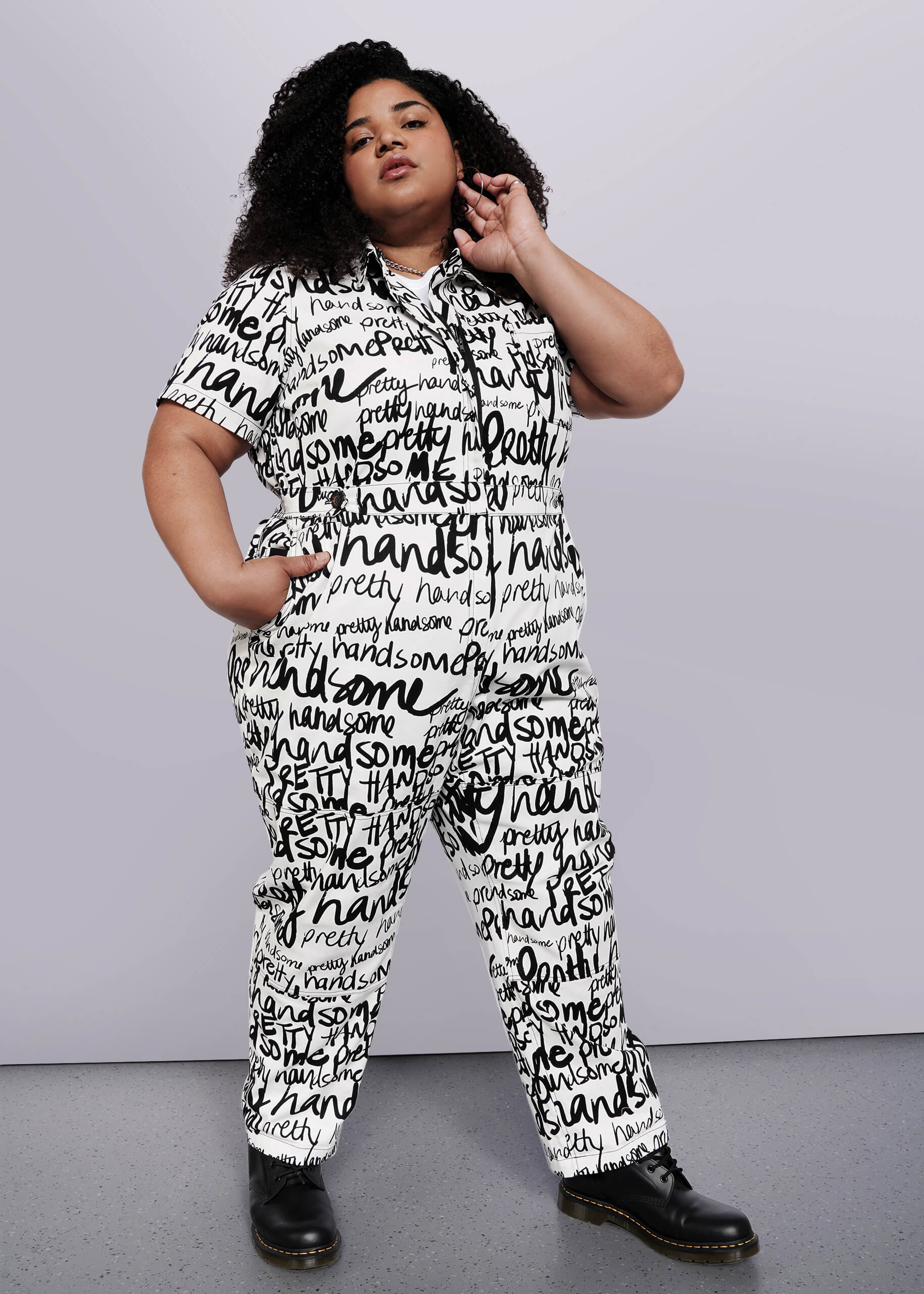 1X=A person with curly hair confidently poses in The Essential High Waisted Coverall, a cotton stretch white jumpsuit featuring bold handwritten black words, paired with black boots against a plain white background.