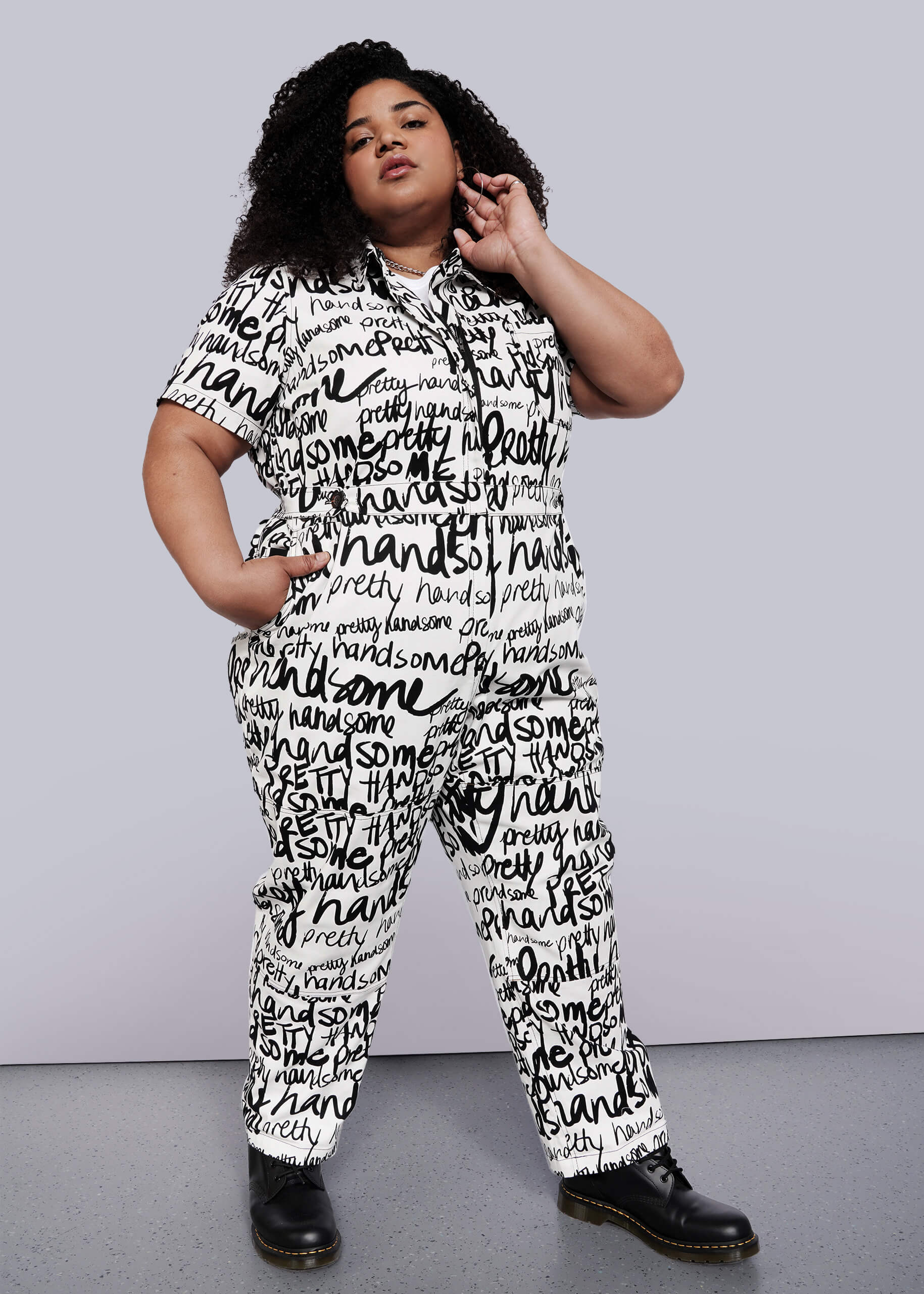 1X=A person with curly hair confidently poses in The Essential High Waisted Coverall, a cotton stretch white jumpsuit featuring bold handwritten black words, paired with black boots against a plain white background.