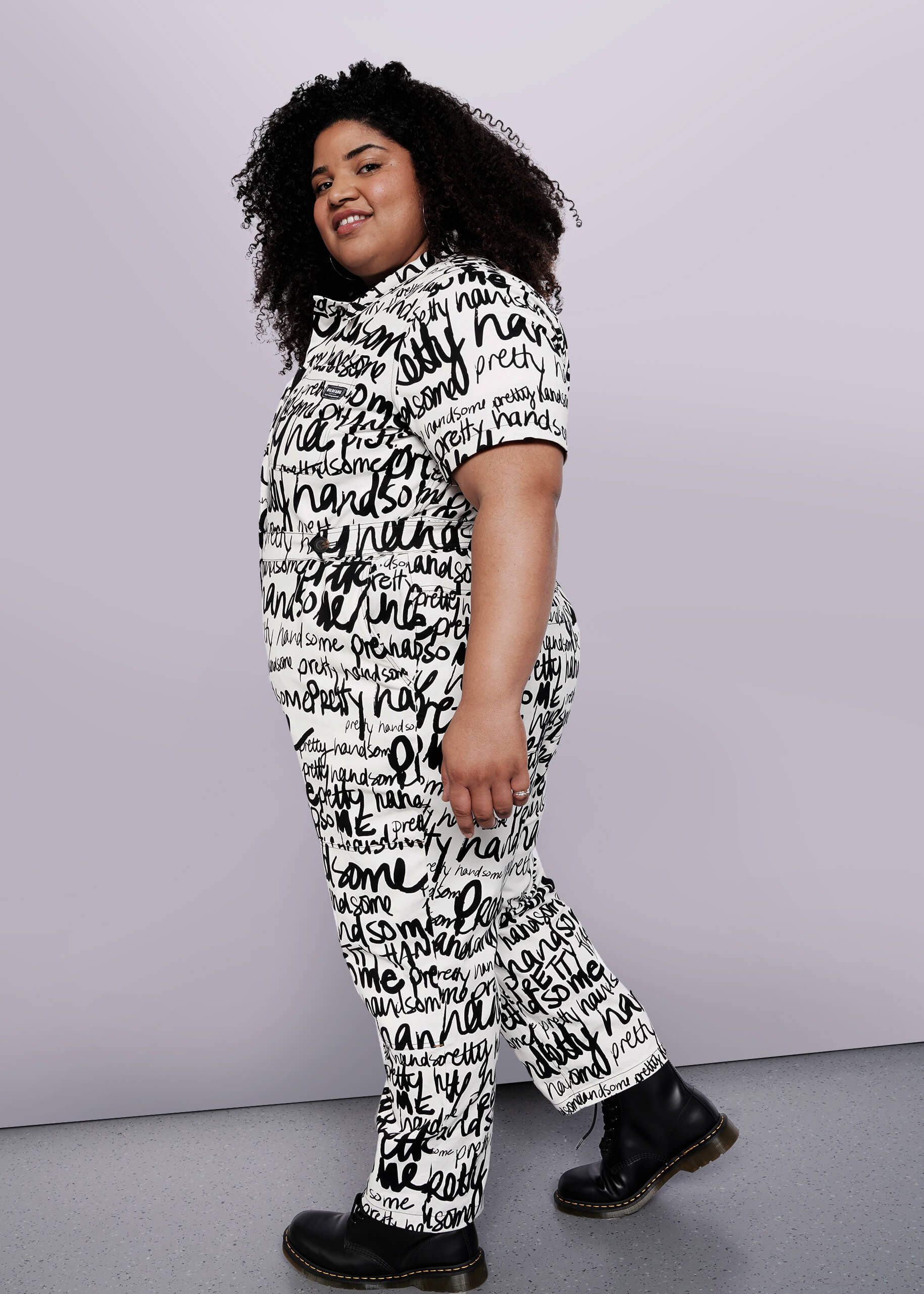 1X=A person with curly hair smiles while posing in The Essential High Waisted Coverall—a white, cotton stretch jumpsuit with deep pockets and black handwritten words—paired with black boots, against a plain white background.