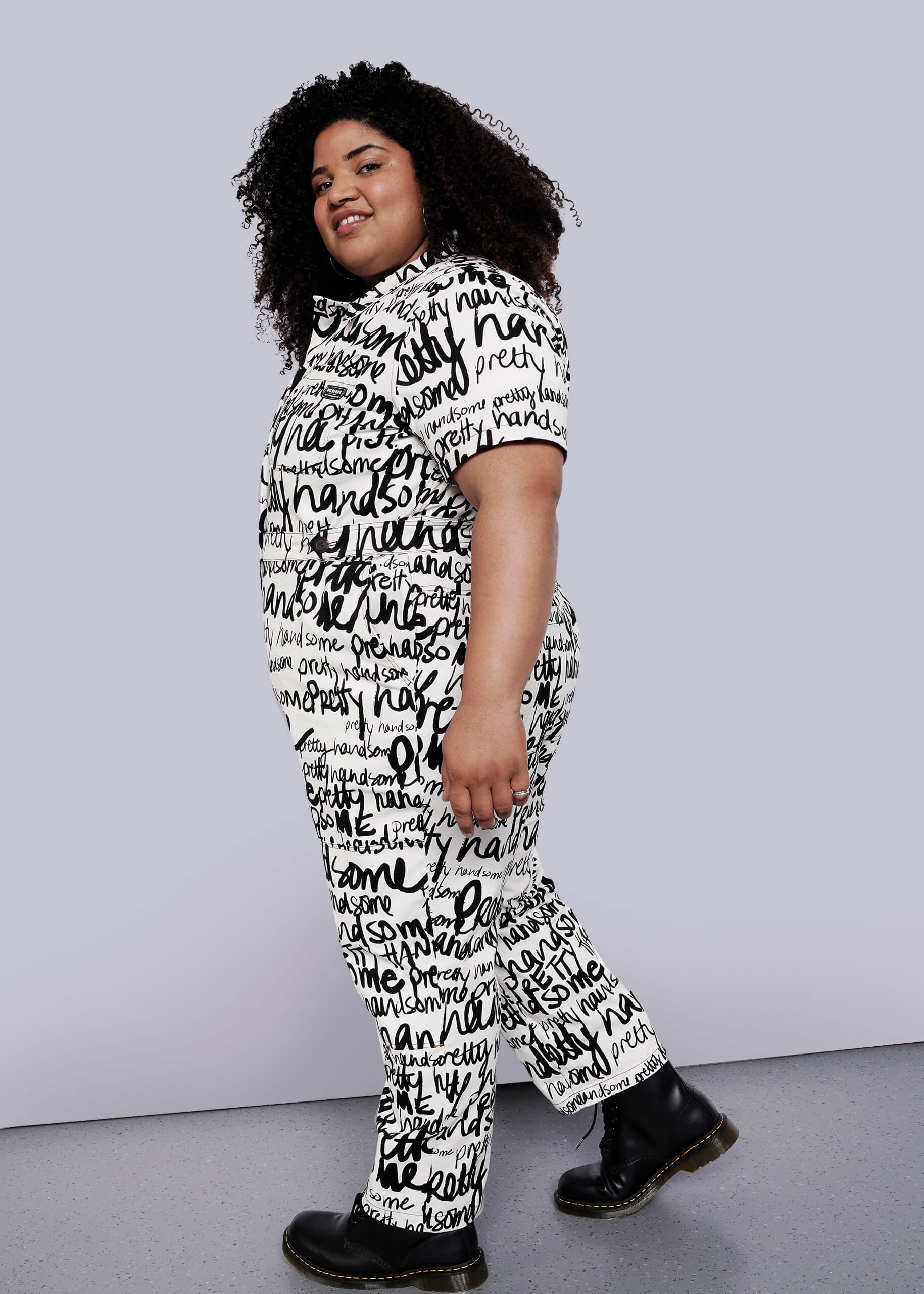 1X=A person with curly hair smiles while posing in The Essential High Waisted Coverall—a white, cotton stretch jumpsuit with deep pockets and black handwritten words—paired with black boots, against a plain white background.