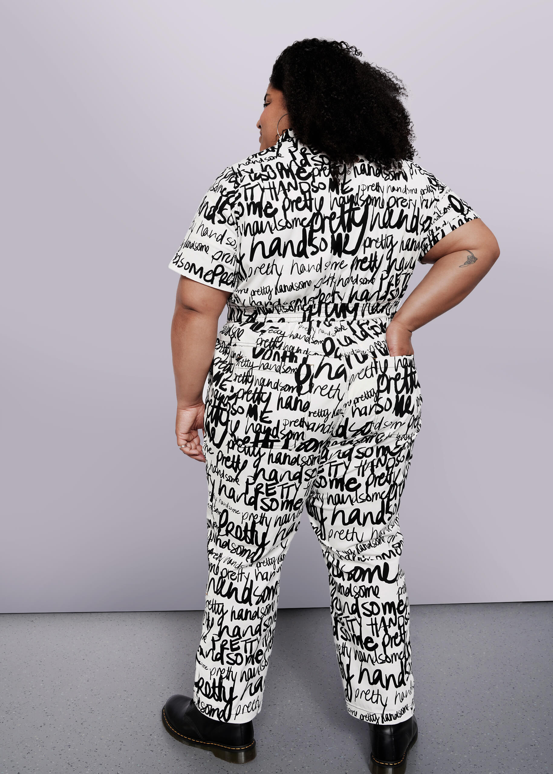 1X=Wearing The Essential High Waisted Coverall, a matching black and white piece covered in handwritten words, a person poses confidently with their back to the camera. Curly hair and black shoes complete this bold look.