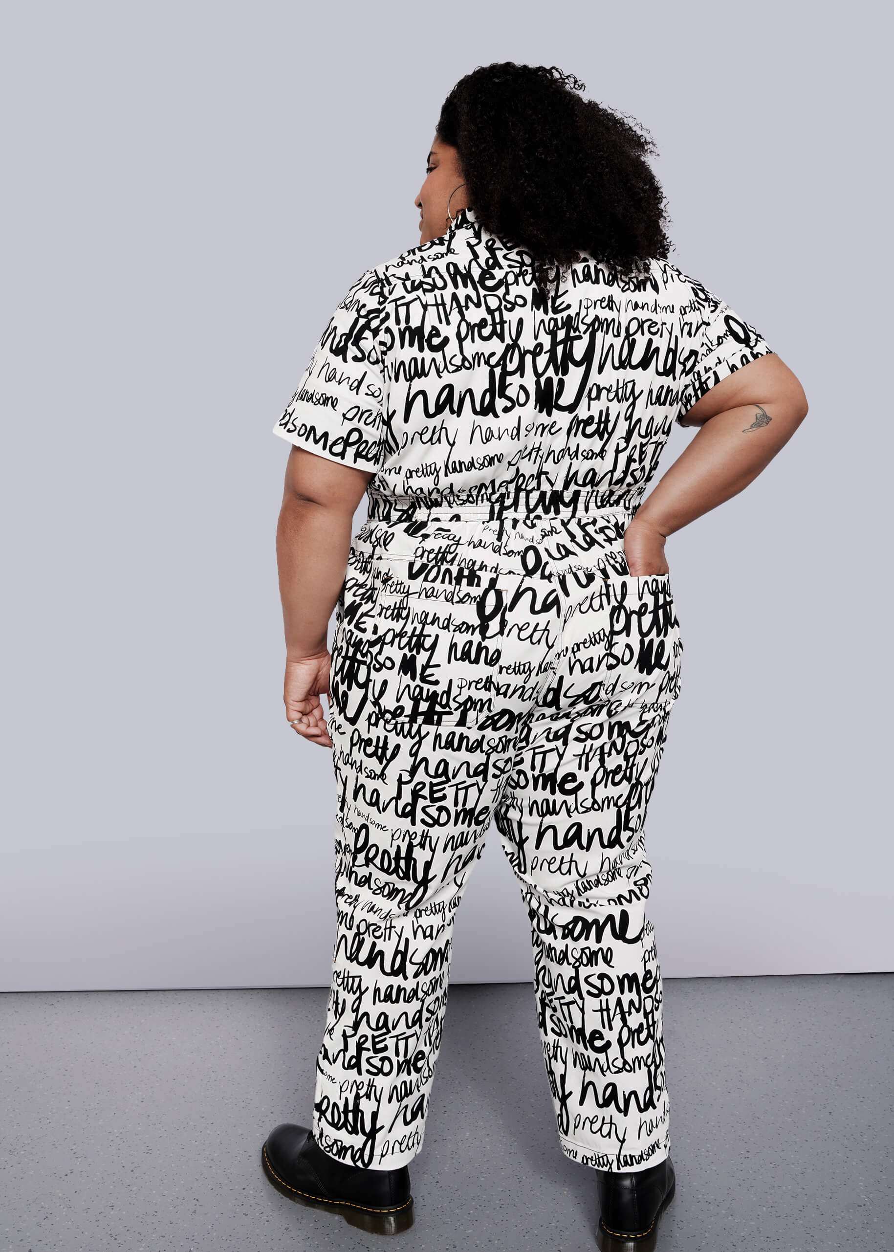 1X=Wearing The Essential High Waisted Coverall, a matching black and white piece covered in handwritten words, a person poses confidently with their back to the camera. Curly hair and black shoes complete this bold look.