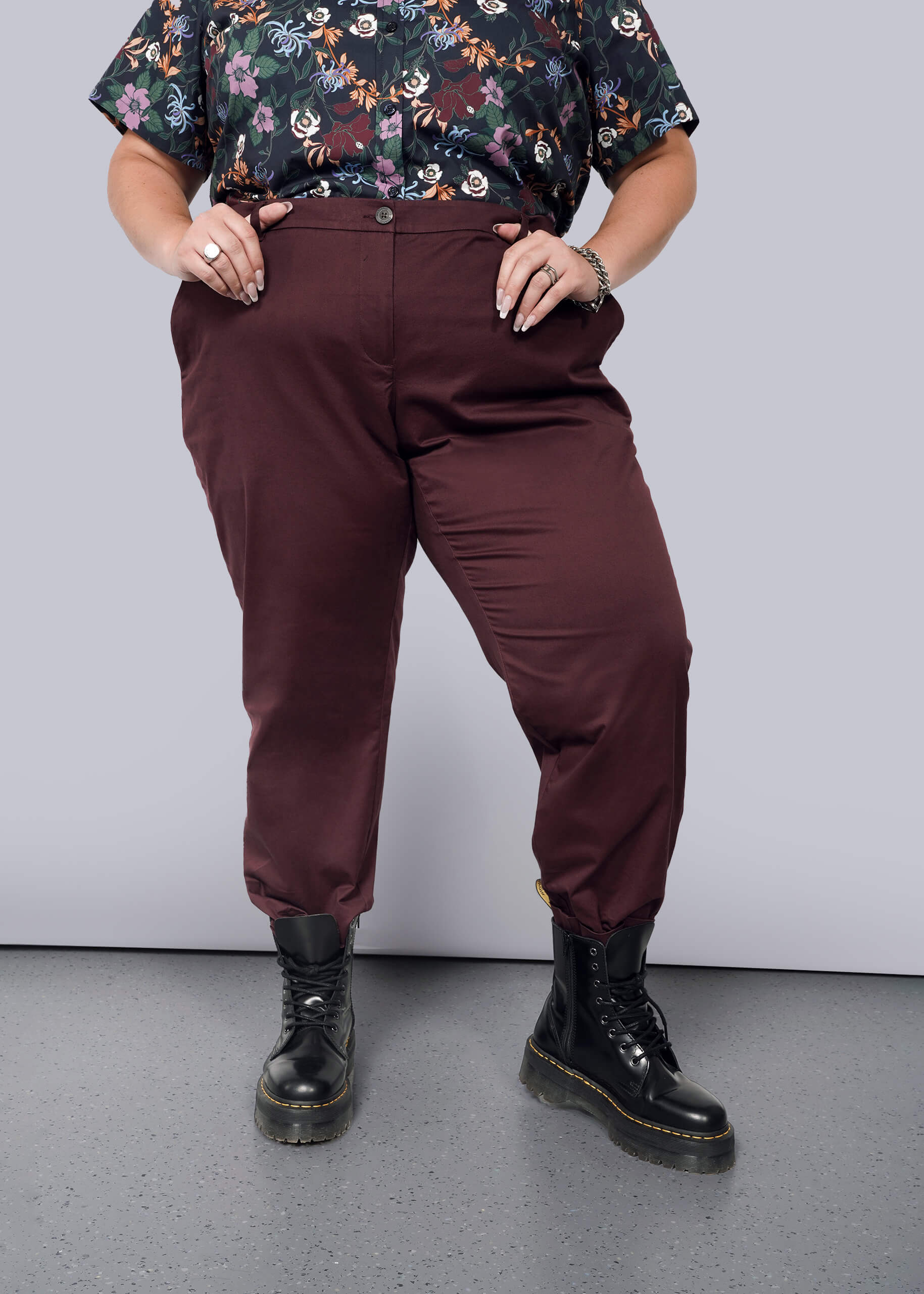 1X=A person in a floral shirt and The Essential Trouser with a burgundy, tapered leg stands with hands in pockets. They wear black boots on a grey speckled floor against a plain white background.