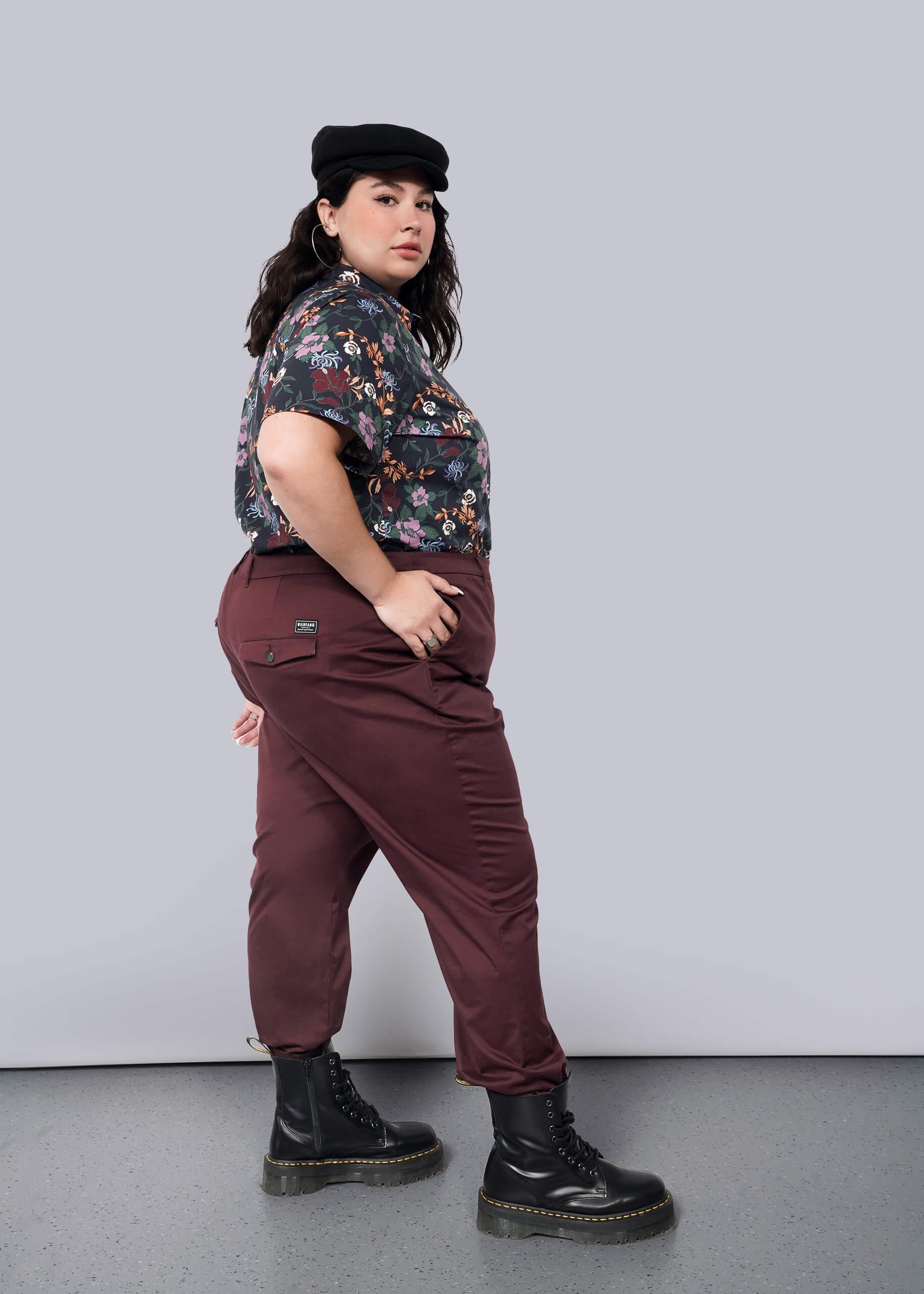 1X=A person stands confidently against a white background, hands in pockets, slightly turned to the side. They wear a black cap, floral shirt, The Essential Trouser in maroon with a tapered leg, and black boots.