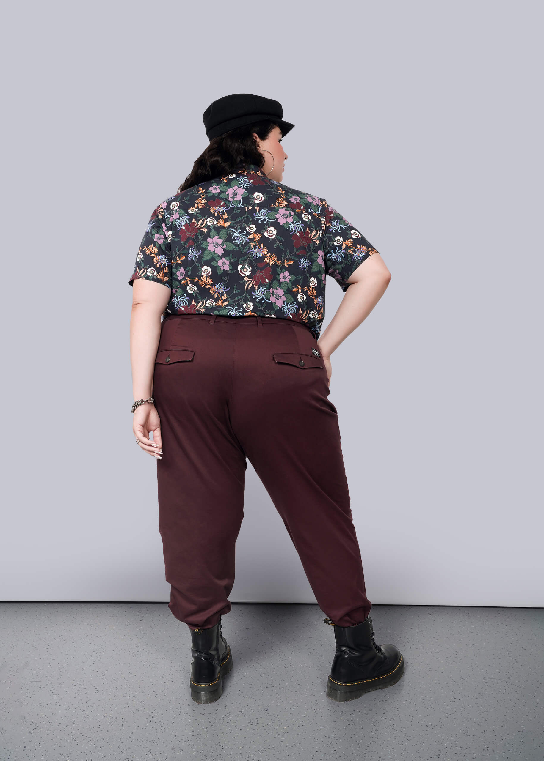 1X=Standing against a plain white background, a person faces away wearing The Essential Trouser in classic maroon with a tapered leg, paired with a floral shirt and black cap. Black boots complete the ensemble as they pose with one hand on their hip.