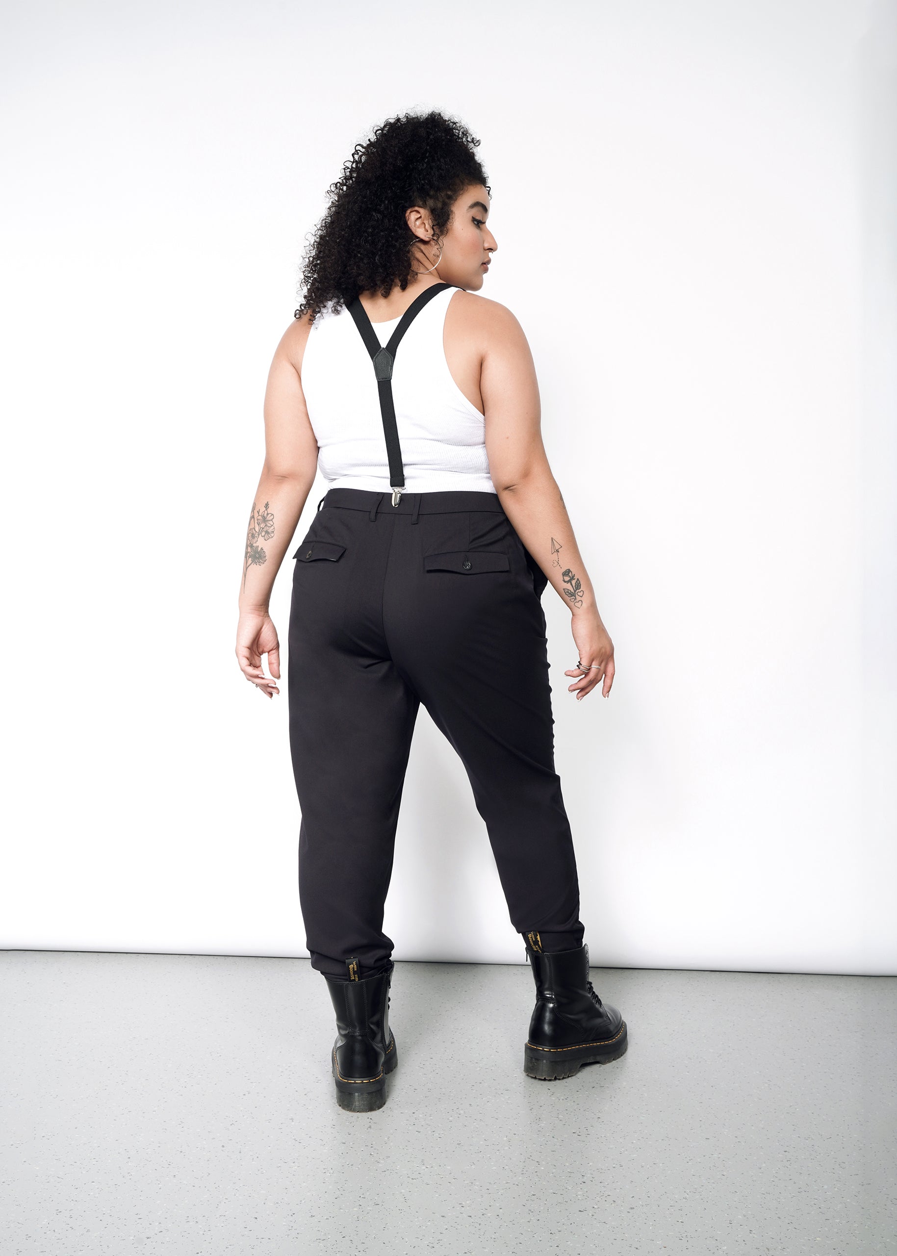 1X=A woman wearing black wildfang pants and a leather jacket. Paired with a white tank, suspenders and doc martens
