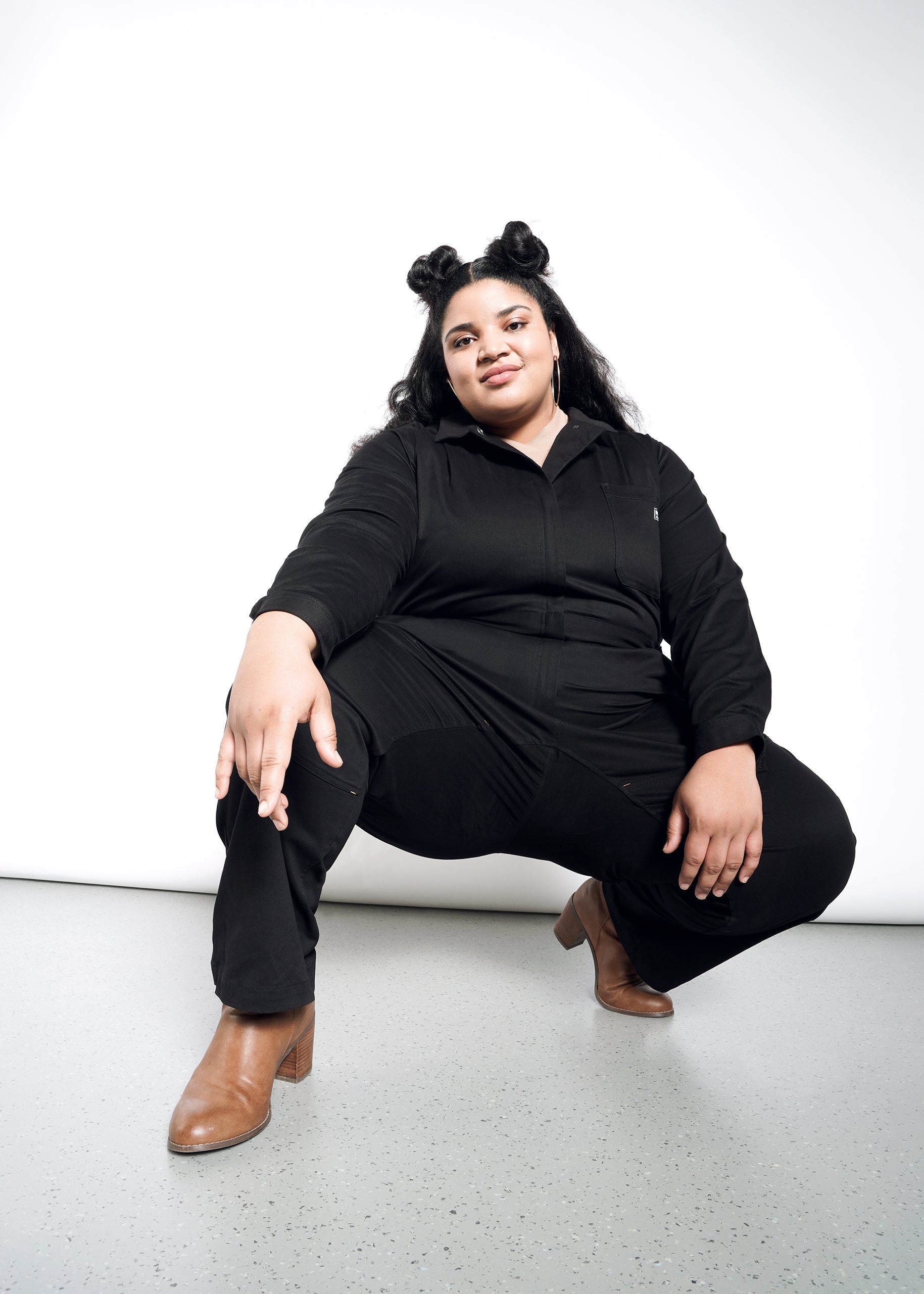 A confident woman with long dark hair in two buns poses in The Essential Long Sleeve High Waisted Coverall and brown heeled boots against a white background, squatting with one hand on her knee and the other reaching forward.