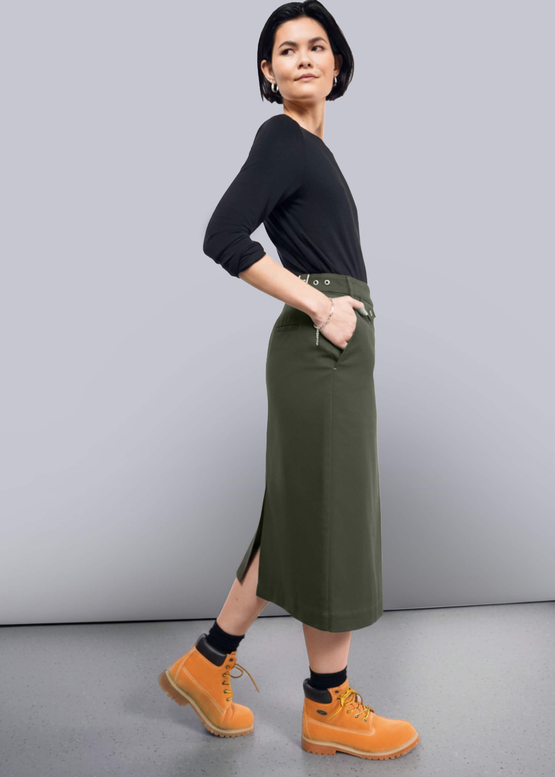 S-=Woman wearing a black top and green skirt with yellow boots on a gray background