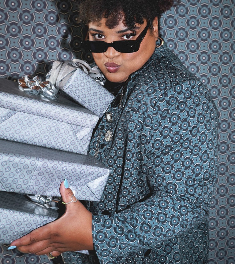 Woman wearing blue patterned suit and black sunglasses holds wrapped gifts in front of blue patterned backdrop