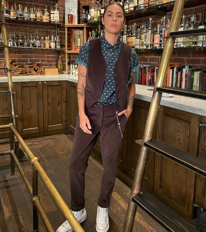 Person stands in bar area wearing a brown corduroy vest and trouser set with a navy blue button up