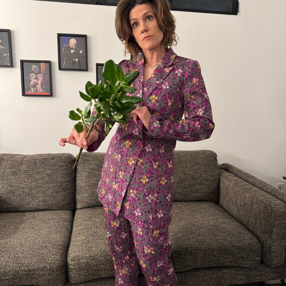 A woman stands in a living room wearing a suit with a pink, purple, and yellow floral pattern. She is holding a green leafy plant in front of a gray textured sofa with framed pictures on the wall.