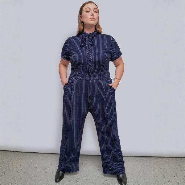Person standing against grey background wearing navy blue pinstripe jumpsuit with black boots.
