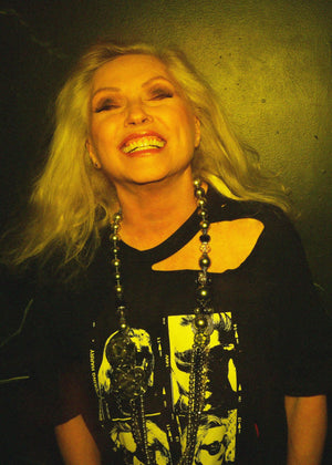 Person with long blonde hair wearing a black shirt with graphic design, against a dark background.