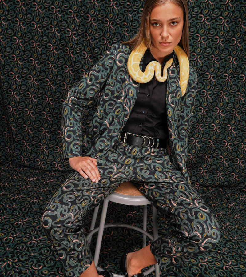 Person on stool wearing Serpentine print suiting looks at camera and has yellow snake around neck. Background is serpentine printed to match suit.