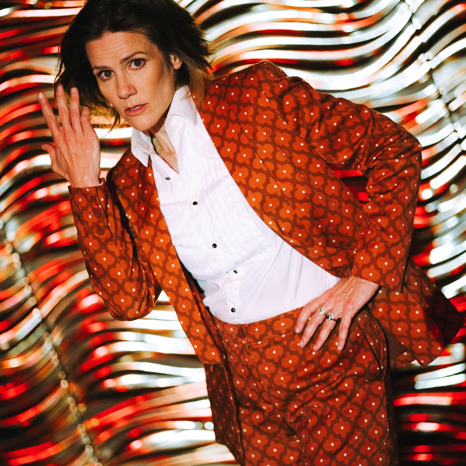 Close-up image of Cameron Esposito wearing a red patterned suit against a swirling red, orange, and white background,