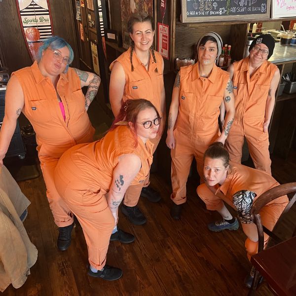 Group of people wearing orange colored sleeveless coveralls standing in restaurant.