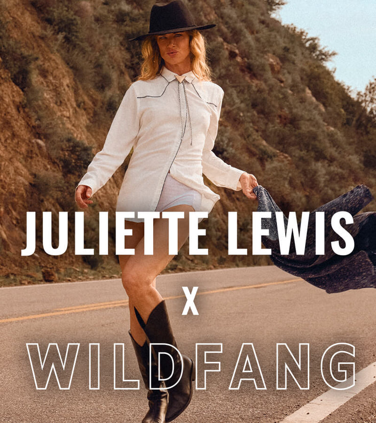 Juliette Lewis stands on road next to mountains wearing cowboy boots, white shorts, a long sleeve white button up, and a black hat. Logo overlay on image says JULIETTE LEWIS X WILDFANG.