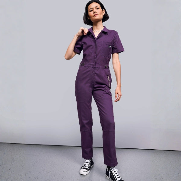 Person standing against grey background wearing purple coverall and black sneakers