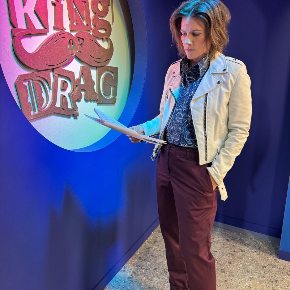 A woman stands indoors wearing a white moto jacket, patterned top, and burgundy pants, holding papers in front of a blue wall with a large 'King of Drag' sign.