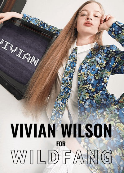 Vivian Wilson sits on floor resting her arm on a vintage TV displaying her name, vivian, on them. She's wearing The Empower Tux Blazer, The Empower Tie, and The Empower Wide Leg Pant all in Carnations Navy, a blue toned floral print. Text overlay reads VIVIAN WILSON FOR WILDFANG.