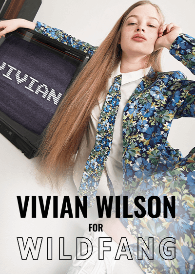 Vivian Wilson sits on floor resting her arm on a vintage TV displaying her name, vivian, on them. She's wearing The Empower Tux Blazer, The Empower Tie, and The Empower Wide Leg Pant all in Carnations Navy, a blue toned floral print. Text overlay reads VIVIAN WILSON FOR WILDFANG.