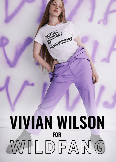 Vivian Wilson stands in front of white backdrop with her name spray painted on it. She wears The Vivian Wilson for Wildfang Tee, which says EXISTING SHOULDN'T BE REVOLUTIONARY, and The Essential High Waisted Coverall in lilac. Text overlay on the image says VIVIAN WILSON FOR WILDFANG.