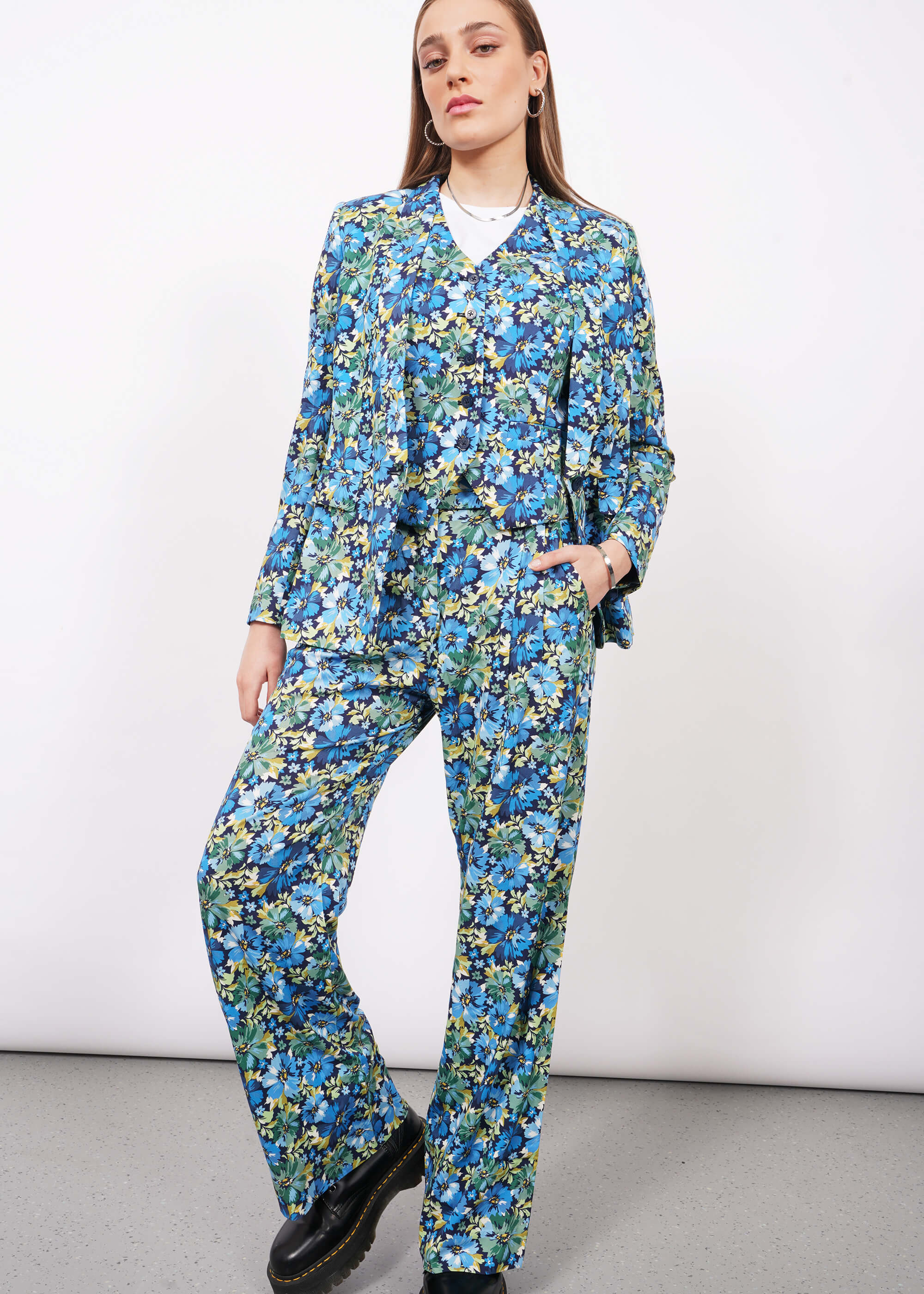 Person with long blonde hair wears The Empower Vest, The Empower Tux Blazer, and The Empower Wide Leg Pant in Carnation Navy, which is a blue toned floral pattern, with black boots.