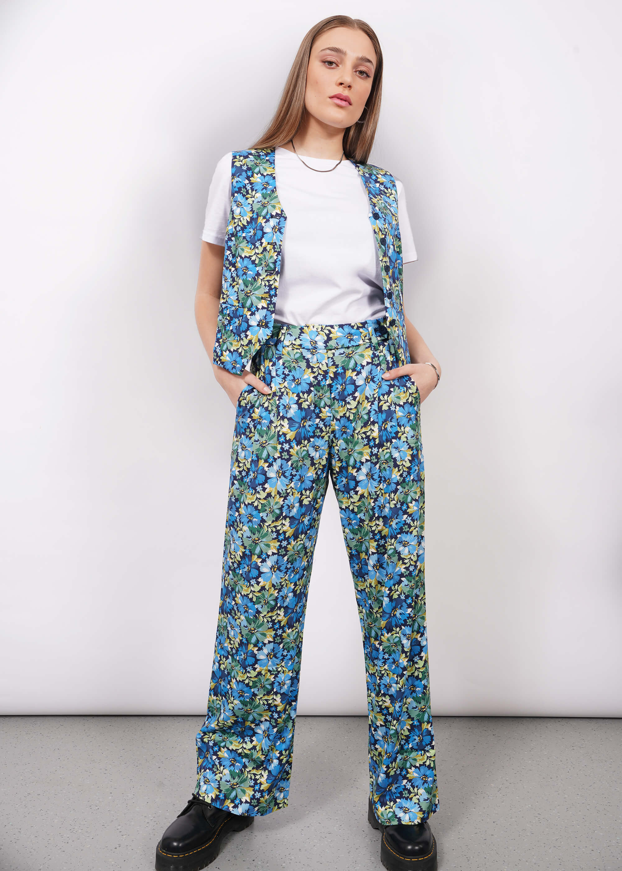 Person with long blonde hair wears a white tshirt, The Empower Vest, and The Empower Wide Leg Trouser both in Carnation Navy, which is a blue toned floral pattern, with black boots.