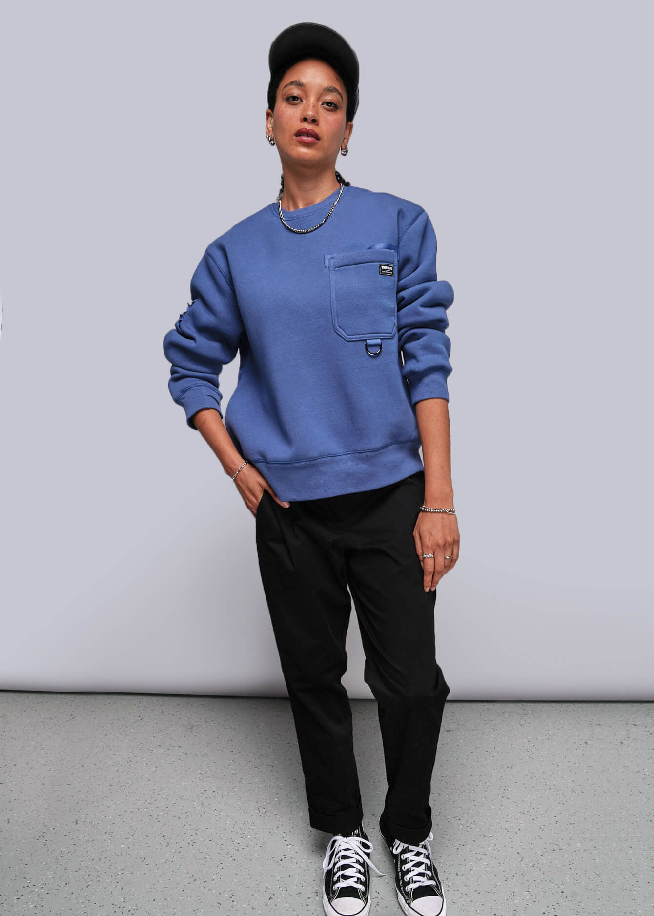 S=Person wearing a blue sweatshirt with a pocket, black pants, and black and white sneakers on a plain background