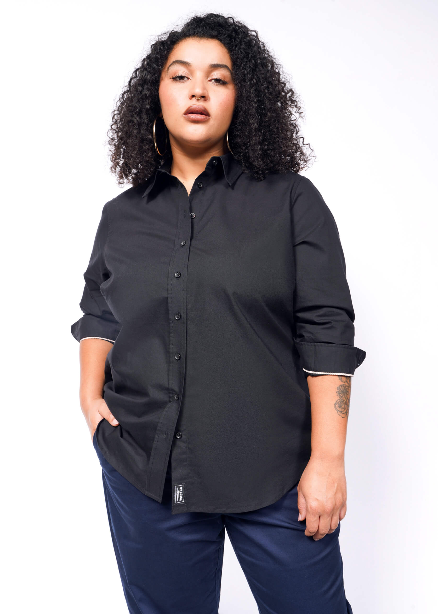 1X=A curly-haired person in The Essential Long Sleeve Oxford Button Up, a tailored fit, black, 100% cotton shirt, pairs it with dark pants and stands with one hand in their pocket.