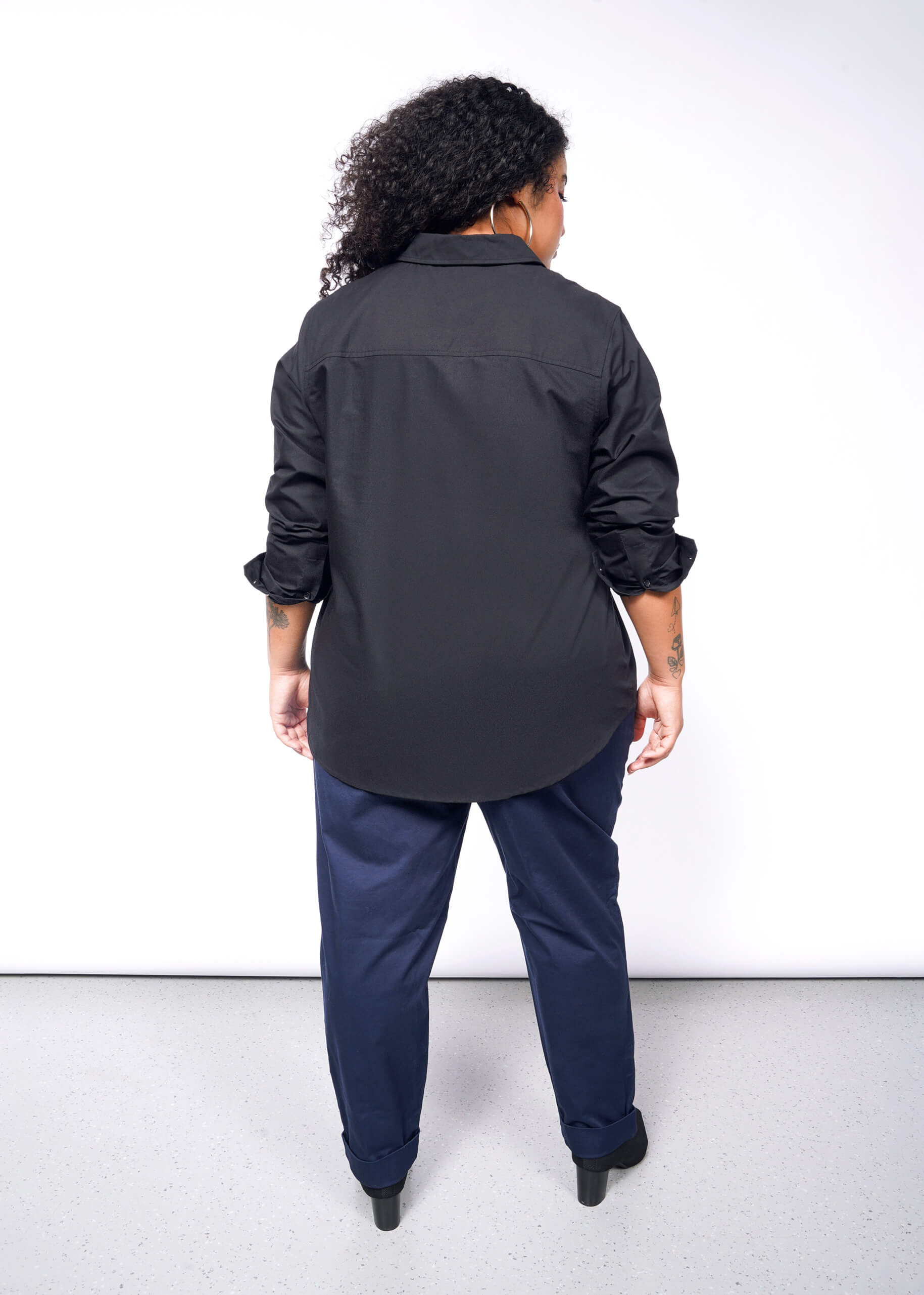 1X=A person with curly hair stands against a white background, wearing The Essential Long Sleeve Oxford Button Up with rolled-up sleeves, navy blue pants, and black shoes. Their tattooed forearms add a distinctive touch to the look.