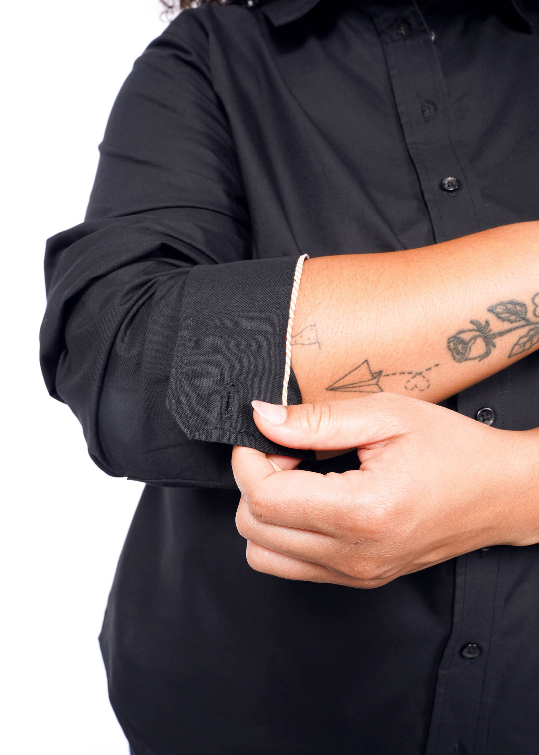 1X=Wearing The Essential Long Sleeve Oxford Button Up, a 100% cotton tailored-fit shirt, a person rolls up their sleeves, exposing forearm tattoos of a paper plane, heart, and rose, while using their right hand to adjust them.