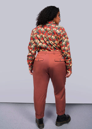 1X=A person with curly hair stands with their back to the camera, showcasing The Essential Trouser in a classic rust color and relaxed mid-rise fit, paired with a floral shirt. Black shoes complete this timeless ensemble against a plain white backdrop.