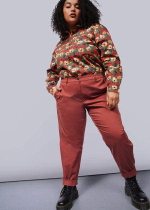 1X=A person with curly hair wears a colorful, patterned shirt and The Essential Trouser in rust color with a relaxed mid-rise fit, paired with black boots. This confident ensemble stands out against a plain white background.