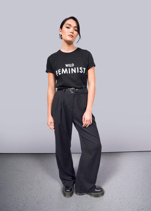 S=Against a white background, someone stands confidently in The Wild Feminist® Tee, a 100% cotton black T-shirt. They pair it with belted black pants and chunky-soled shoes, with hands relaxed at their sides.