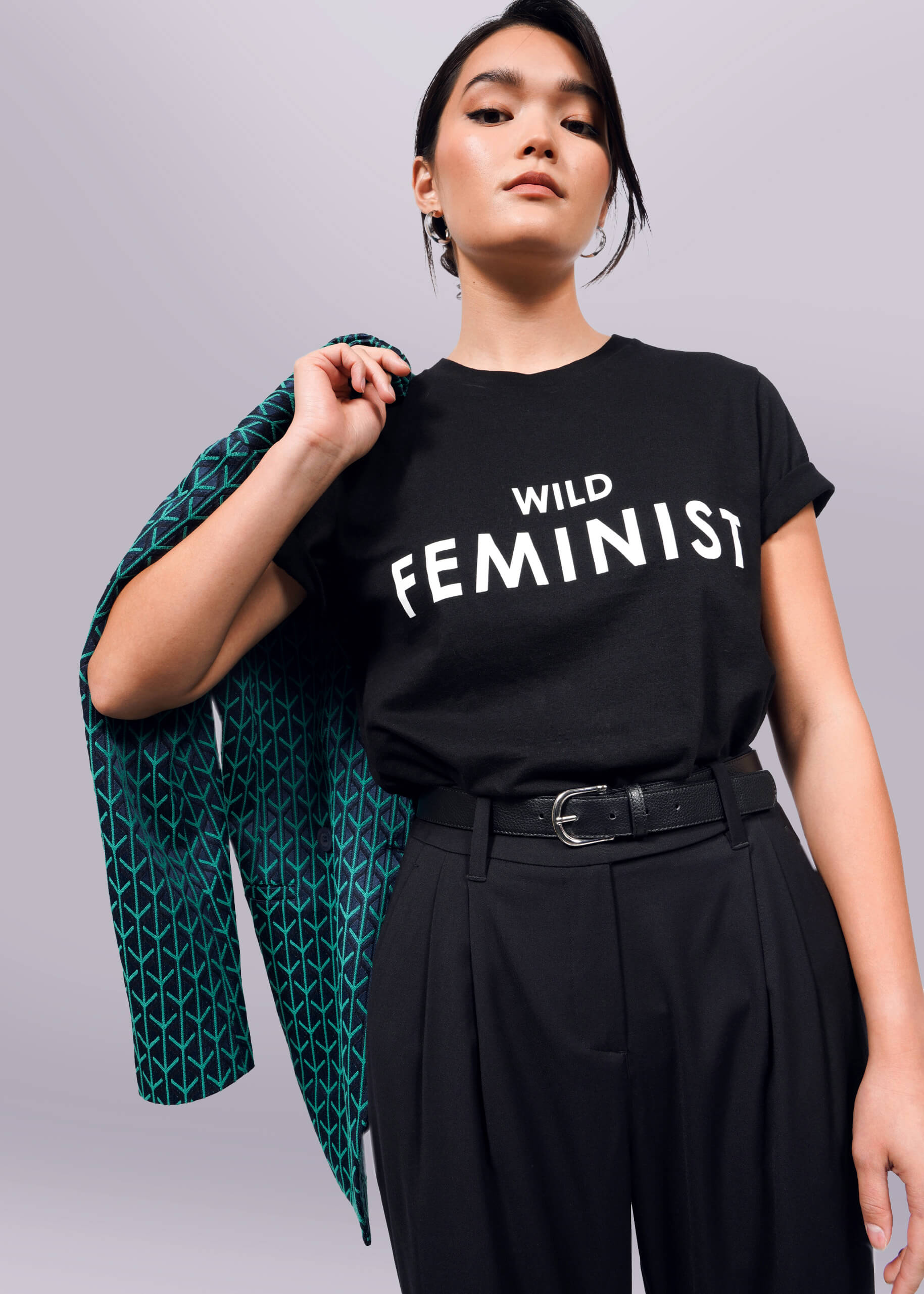 A woman confidently poses in The Wild Feminist® Tee, a black unisex fit T-shirt with WILD FEMINIST in bold white letters, made from pre-shrunk 100% cotton for lasting comfort. She holds a jacket over her shoulder and pairs it with high-waisted black pants against a plain white background.